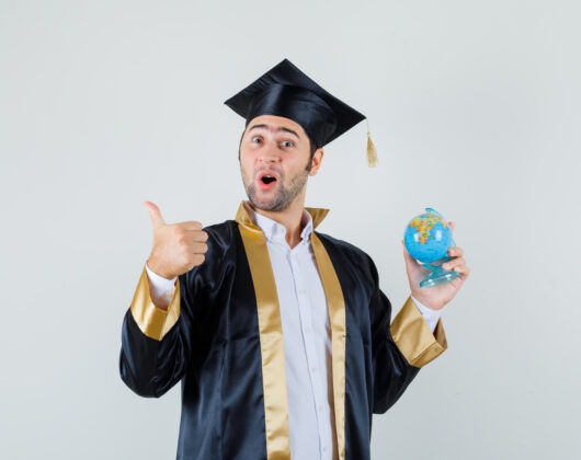 Young man in graduate uniform holding globe in his hand showing thumb up