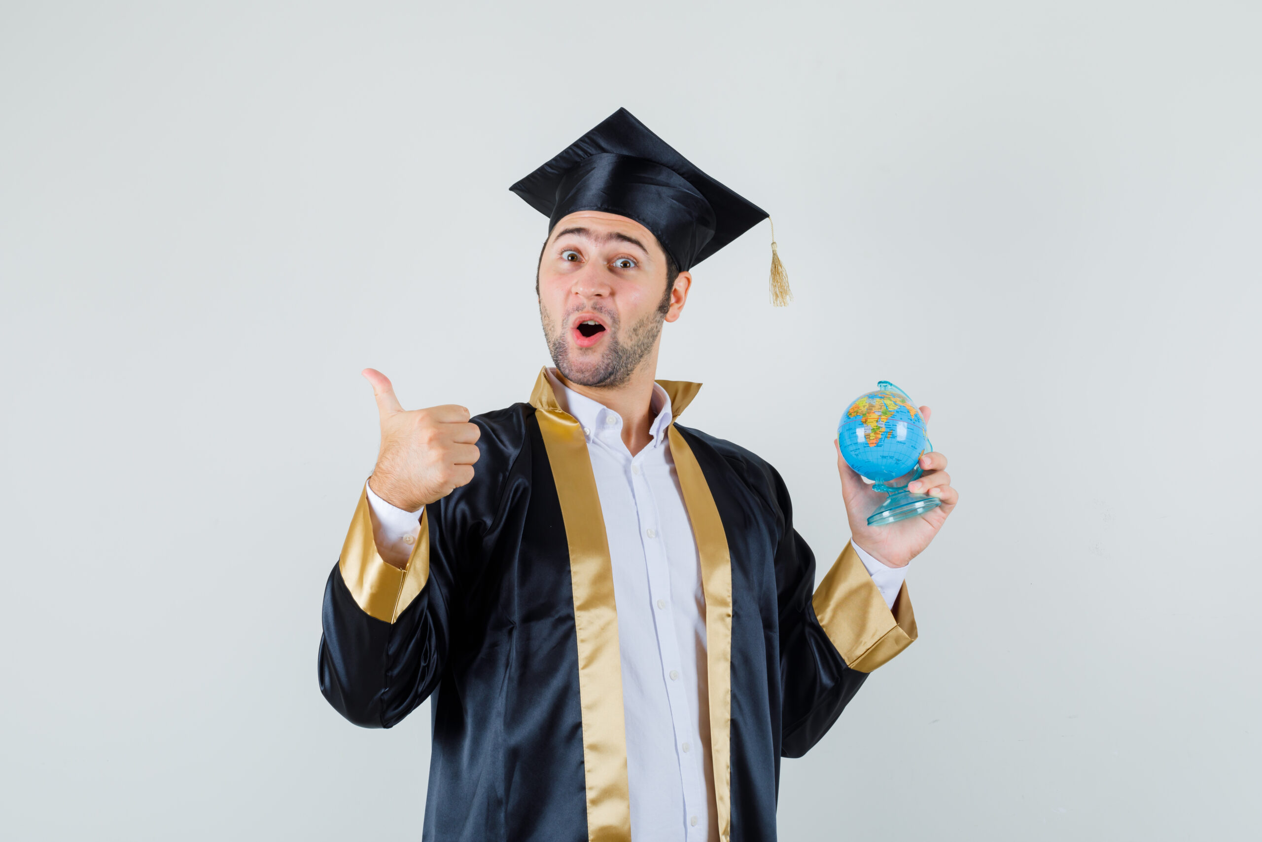Young man in graduate uniform holding globe in his hand showing thumb up
